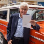 Jay Leno holding signed paper beside orange classic Ford Bronco