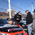 A Driver Standing next to a Car Signing Autographs during a Race Event A Driver Standing next to a Car Signing Autographs during a Race Event